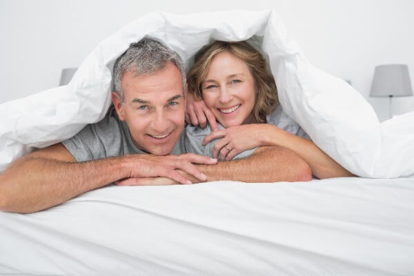 Paar liegt auf dem Bauch im Bett und schaut am Fußende lächelnd unter einer Weißen Bettdecke hervor.