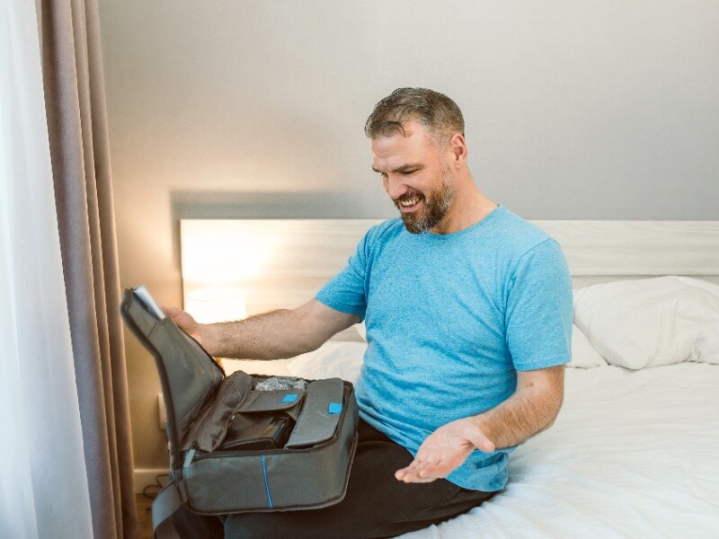 Grauhaariger Mann mit Bart sitzt auf einem Bett und schaut in die Tasche mit einem PAP-Gerät. Er trägt ein türkises T-Shirt.