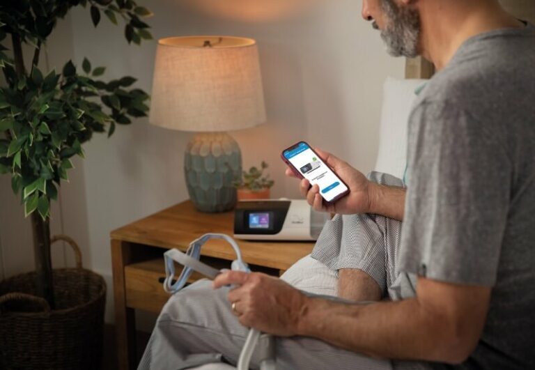 Mann sitzt mit angewinkeltem Bein auf dem Bettrand. er hält ein Handy mit dem Screen einer digitalen Therapiebegleitung in der rechten Hand und eine Beatmungsmaske in der linken.