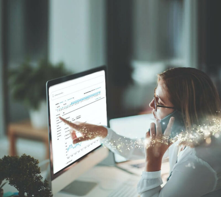 Eine Frau mit Brille sitzt vor einem Monitor, auf dem Auswertungsdiagramme gezeigt werden. Sie hält ein Handy an ihr linkes Ohr und zeigt auf den Bildschirm.