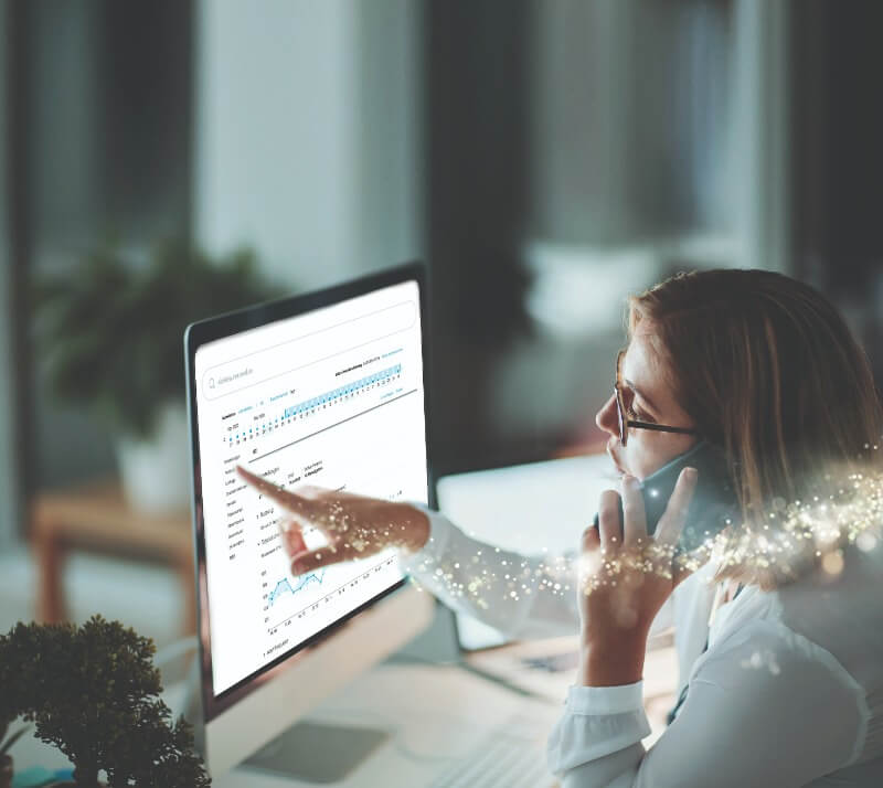Eine Frau mit Brille sitzt vor einem Monitor, auf dem Auswertungsdiagramme gezeigt werden. Sie hält ein Handy an ihr linkes Ohr und zeigt auf den Bildschirm.