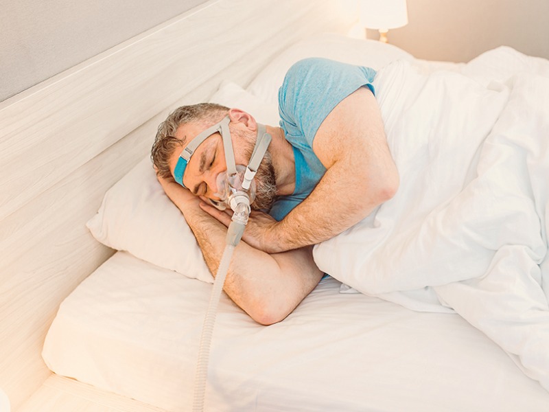 Grauhaariger Mann mit Bart liegt auf der rechten Seite im Bett. Er trägt eine PAP-Maske und hat die Hände unter die Wange geschoben.