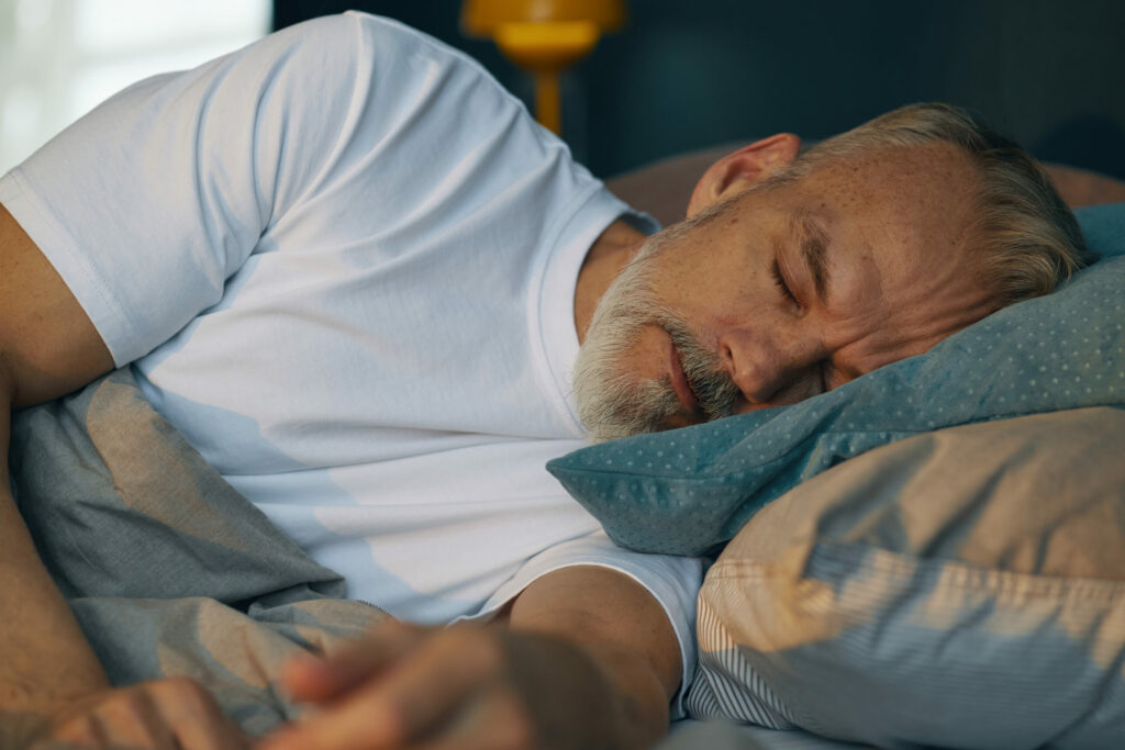 Grauhaariger Mann mit Bart im weißen T-Shirt liegt auf der linken Seite im Bett und schläft friedlich.