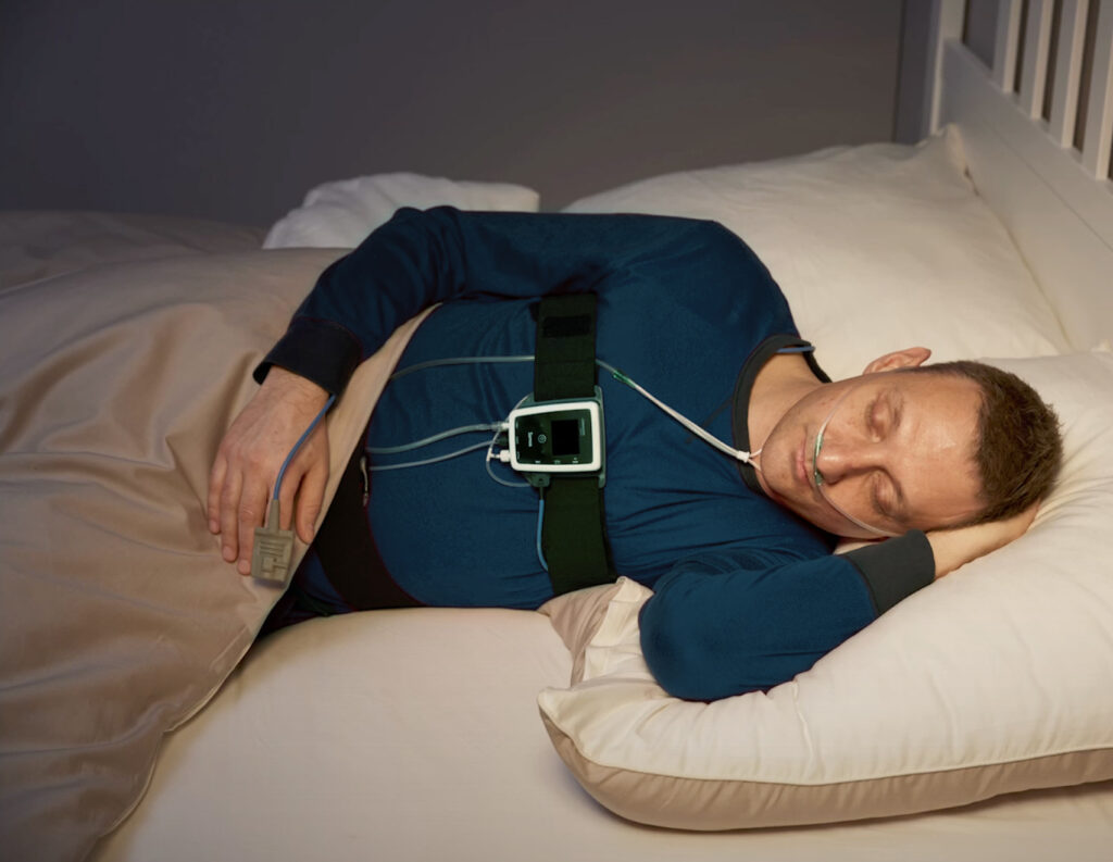 Mann in blauem Pyjama liegt schlafend mit einem Heimpolygraphie-Gerät im Bett.